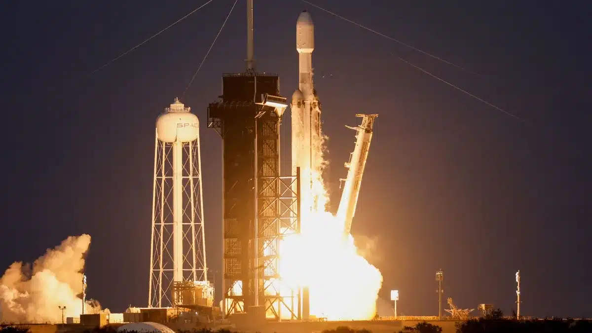 Falcon Heavy da SpaceX decolando à noite no Centro Espacial Kennedy, com três colunas de fogo intensas iluminando o céu e fumaça densa na base da plataforma.